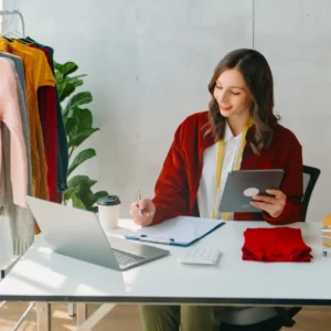 A woman sat at a desk in a stock room writing on a clipboard and holding a tablet