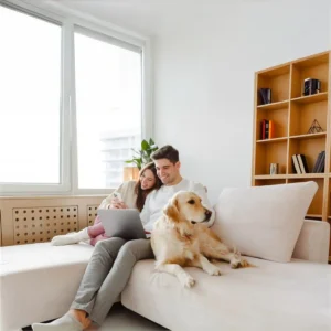 Young couple sat on a sofa using a laptop and phone with their golden retriever dog