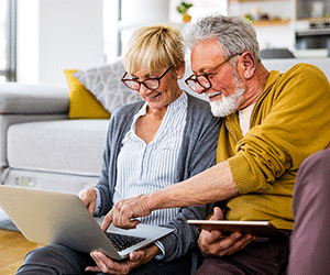 An elderly couple using a laptop and a tablet sat in their living room at home