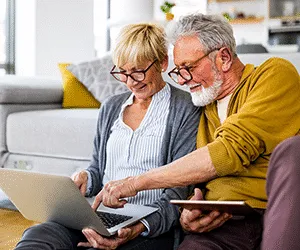 An elderly couple using a laptop and a tablet sat in their living room at home